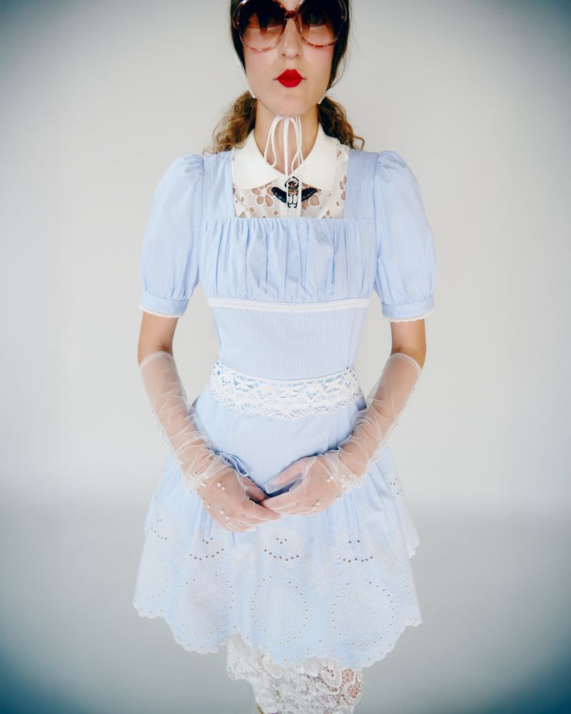 Woman in a blue eyelet dress with lace details, accessorized with sheer gloves and red sunglasses, standing in front of a grey studio background.