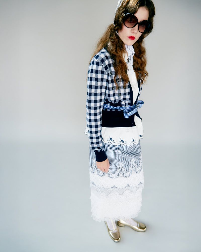 A young woman poses in a blue and white gingham skirt and jacket, accessorized with a white headscarf and oversized sunglasses, against a gray background.