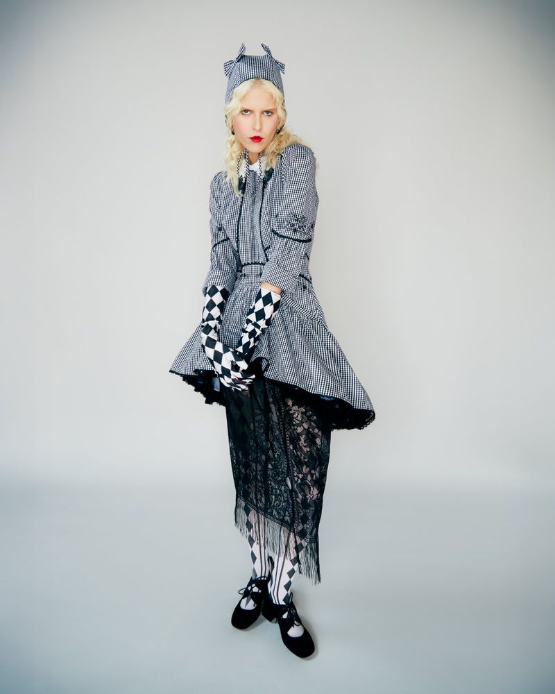 Full shot of a female model with blonde curly hair, in a grey and black checkered dress and hat, plus black-and-white gloves and tights.