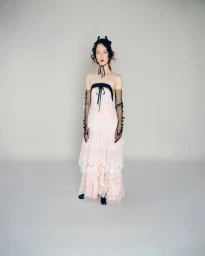 Model standing in a tiered pale pink tulle dress and sheer black opera gloves against a studio background for Bora Aksu S/S 26.