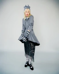Model posing in a grey checkered dress, harlequin print gloves, and black lace skirt against a neutral studio backdrop.