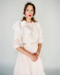 Model wearing a pale pink dress with a large shoulder bow and lace bonnet against a plain light background