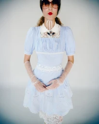 Model in light blue dress with white lace collar and sheer gloves standing against a seamless white background.