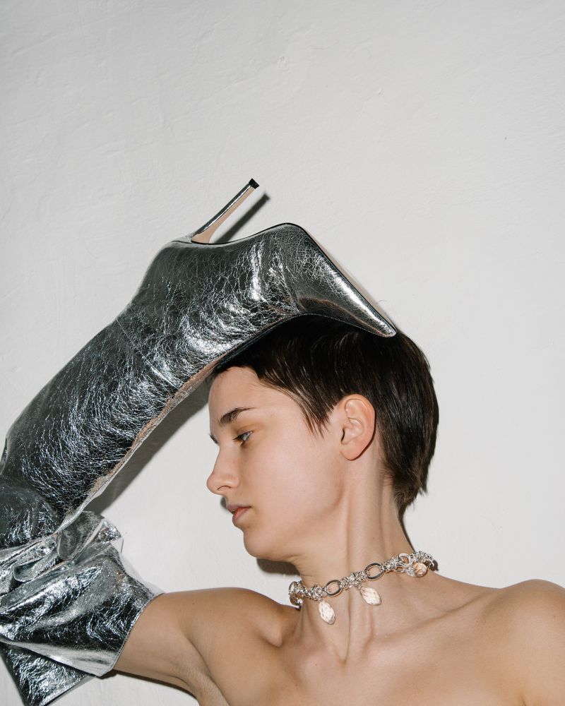 Woman wearing silver glove and boot on head, jewelry, against a plain wall. Fashion and unconventional style.
