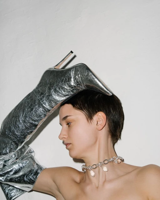 Woman wearing silver glove and boot on head, jewelry, against a plain wall. Fashion and unconventional style.