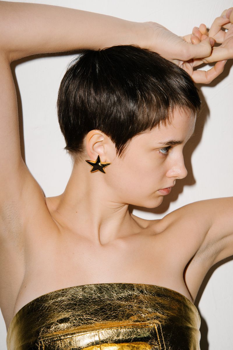 Glamour shot of a young adult woman with short dark hair wearing large star earrings and a golden strapless top against a white backdrop.