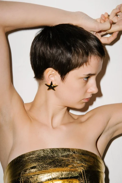 Glamour shot of a young adult woman with short dark hair wearing large star earrings and a golden strapless top against a white backdrop.