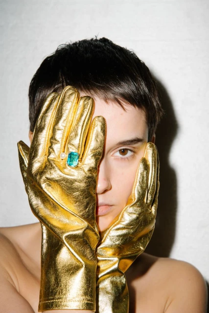 Close-up of a woman with short dark hair wearing gold gloves with a large gemstone ring, posing in an indoor studio setting with neutral background.