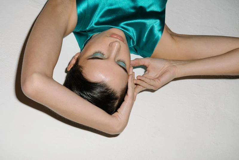 Elegant woman with blue eyeshadow poses with hands near face while lying on white surface in fashion glamour beauty studio shot.