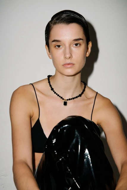 Portrait of a young woman with slicked-back hair wearing a black top and beaded necklace with a dark pendant, posing in front of a white wall.