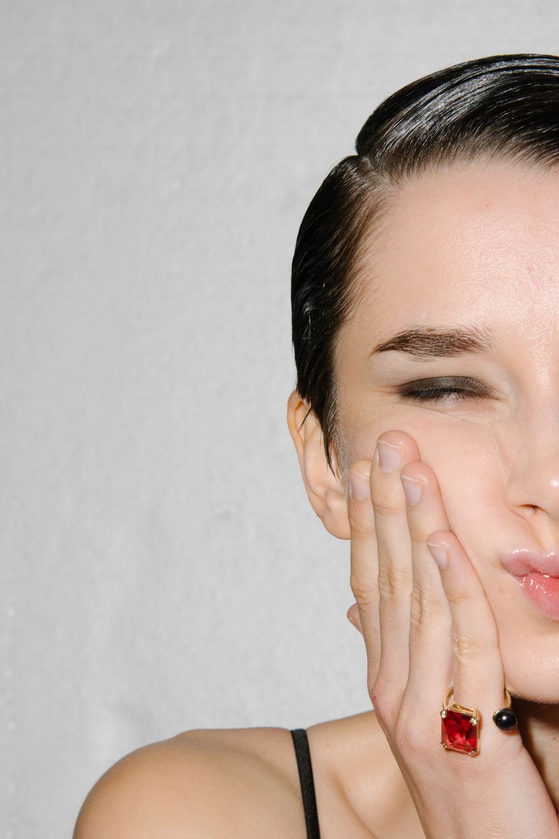 Close-up of a woman's face with slicked-back hair and a large ruby ring. Her hand is against her face, highlighting her makeup.