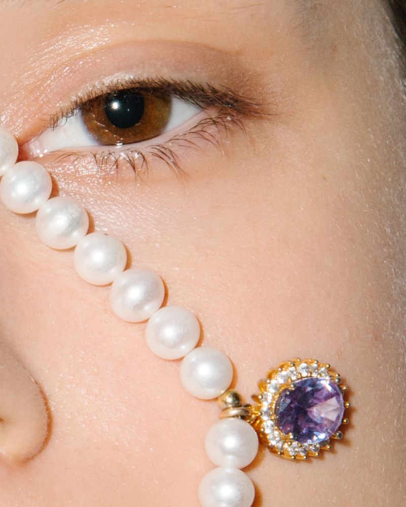 Close-up on a fair-skinned woman's face featuring a brown eye, adorned with a string of pearls, and a purple gemstone earring.