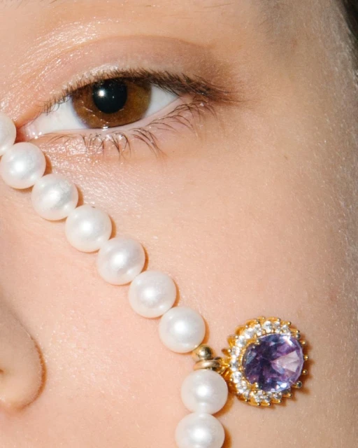 Close-up on a fair-skinned woman's face featuring a brown eye, adorned with a string of pearls, and a purple gemstone earring.