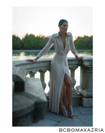 Woman in elegant dress posing on a stone balcony with a view of a pond and trees, under a sky at dusk, fashion editorial shot.