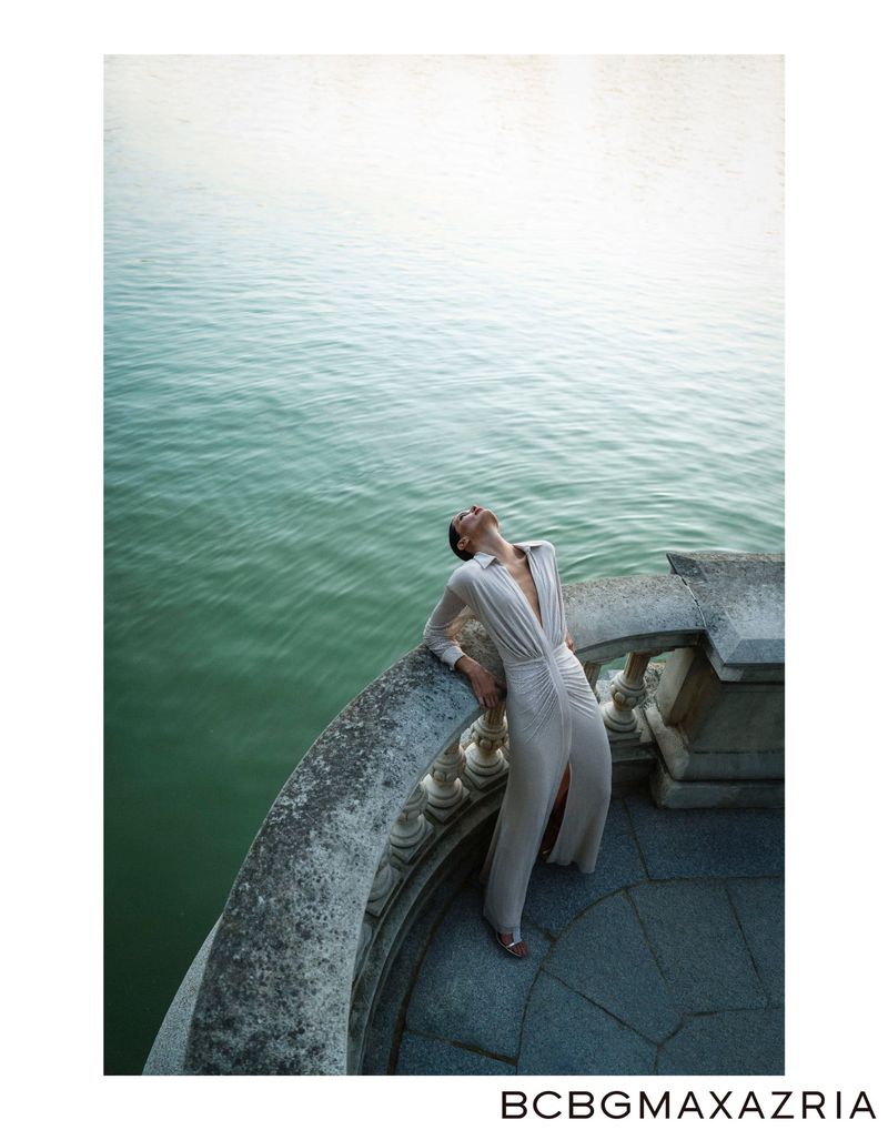 A woman in a white dress leans back against a stone railing with a body of water behind her in an outdoor fashion shot.