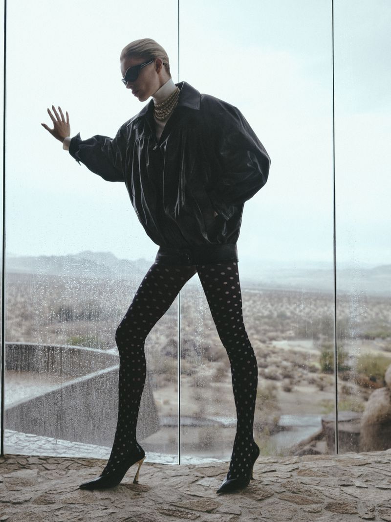 A woman in a black jacket, patterned tights, and heels poses in front of a glass wall with a desert landscape background.