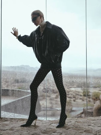 A woman in a black jacket, patterned tights, and heels poses in front of a glass wall with a desert landscape background.