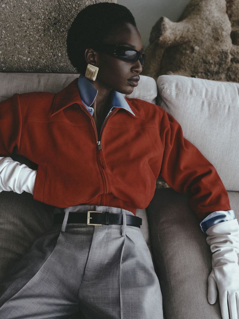 Model Zayna Cisse wearing a rust zip-up jacket, grey pleated trousers, white leather gloves, and sunglasses while seated on a couch.
