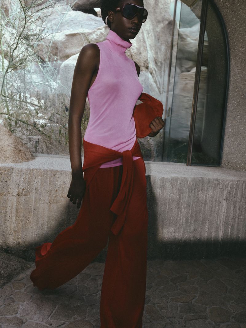 Fashionable woman in pink top and red pants poses outdoors against rocky wall. She carries a red clutch, wearing oversized sunglasses.