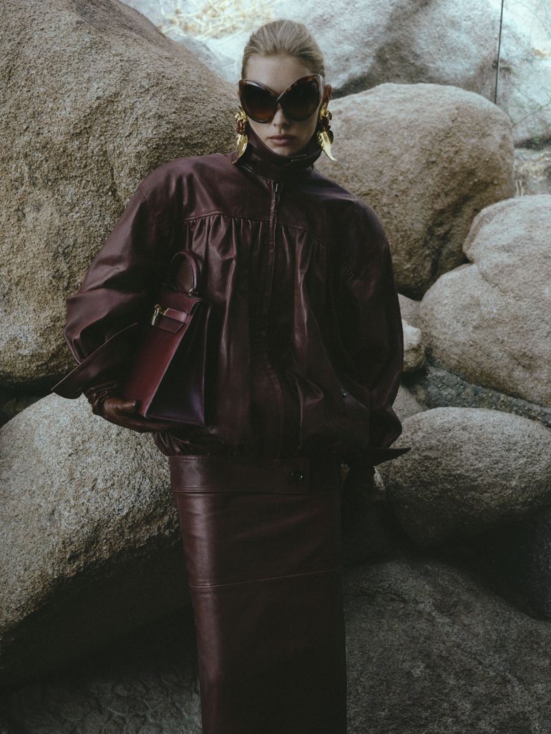 Woman poses in a burgundy leather jacket and pencil skirt with tan rocks, wearing sunglasses and gold earrings in a fashion shot.