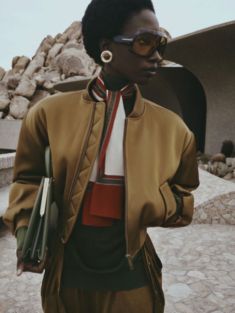 A fashionable Black woman wearing sunglasses, jacket, and carrying a purse, stands against a unique architectural backdrop with rock formations.