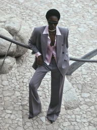 A model wearing a grey pantsuit and pink silk shirt stands outdoors on a stone-paved stairway.