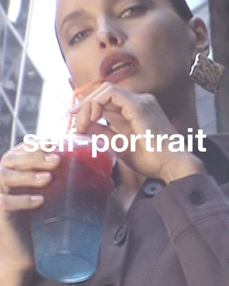 Close-up of a woman outdoors drinking a blue and red beverage with a straw; she wears gold earrings and a brown jacket.
