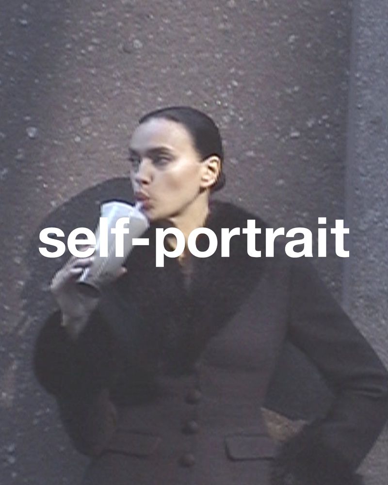 A young adult woman with dark hair in a sleek updo sips from a straw in a clear cup while posing against a textured wall.