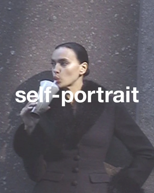 A young adult woman with dark hair in a sleek updo sips from a straw in a clear cup while posing against a textured wall.