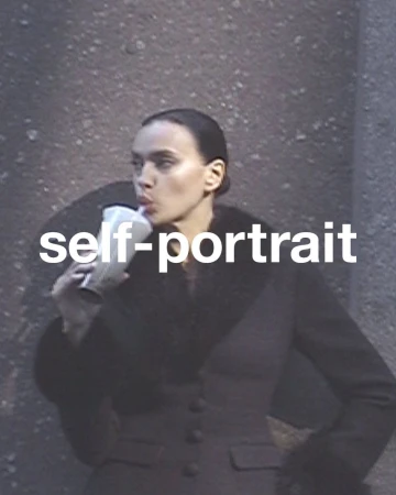 A young adult woman with dark hair in a sleek updo sips from a straw in a clear cup while posing against a textured wall.