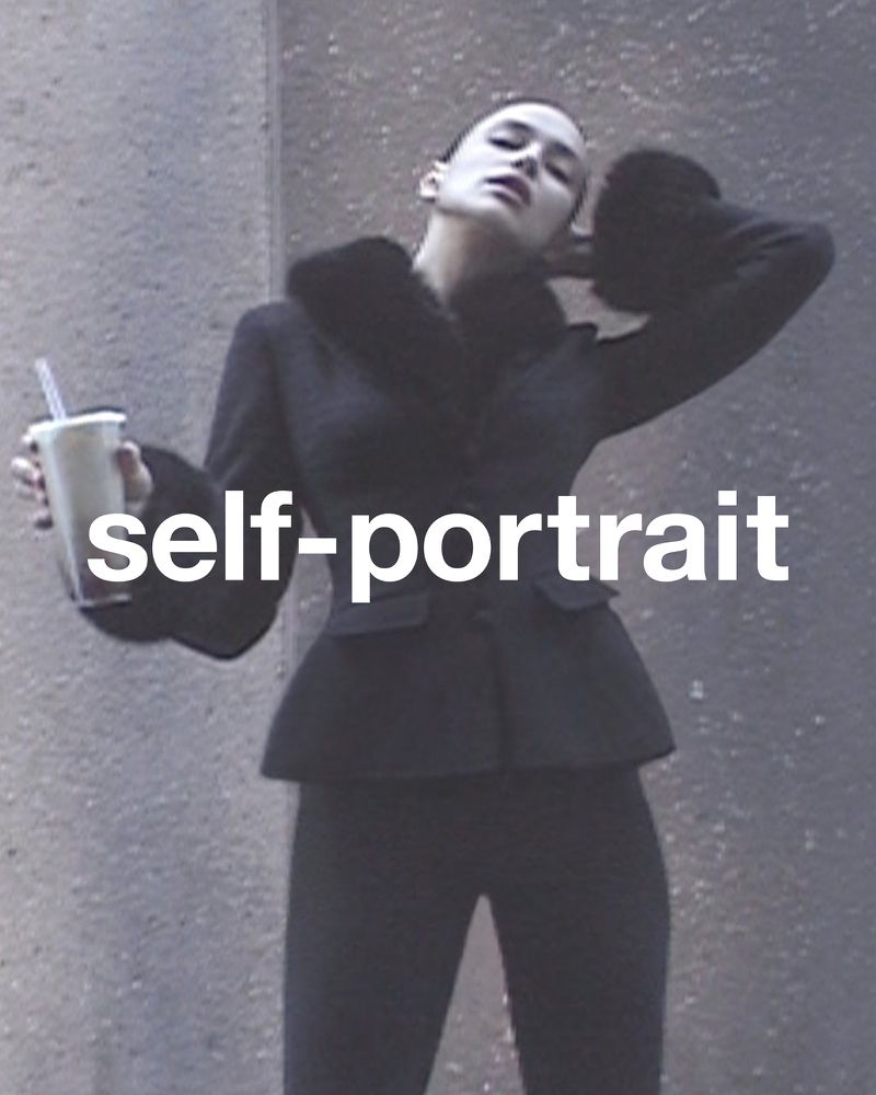 A woman poses with a drink outdoors, wearing a black jacket and pants, captured in a portrait with a neutral expression and urban setting.