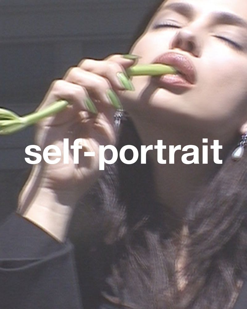 Woman posing in a self-portrait, holding and eating green vegetables, wearing black blazer and silver earrings, against a blurry background.