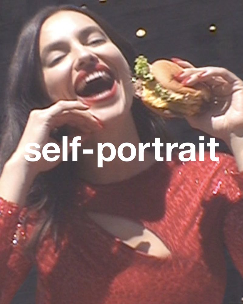 A woman with long, dark hair and red lipstick wearing a red sequin shirt, holds a burger in her hand with a big smile on her face.