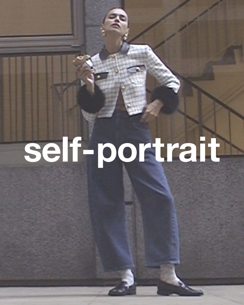 Woman poses in a tweed jacket, jeans, and loafers on a city street, with a staircase visible in the background during the daytime.