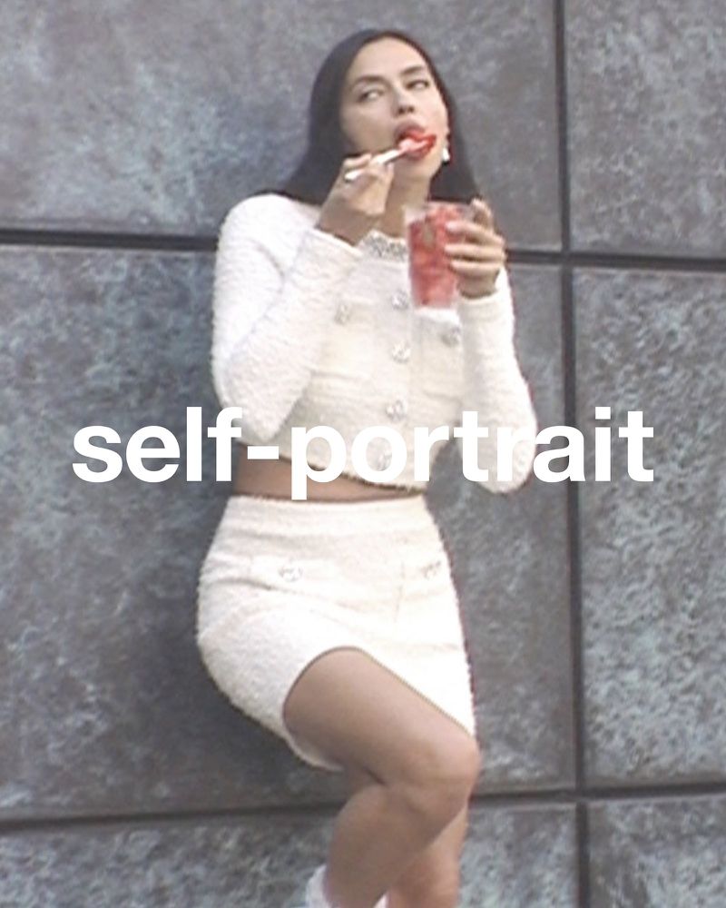 A woman in a white two-piece outfit eats from a cup with a spoon against a textured wall. The photo is a medium shot.
