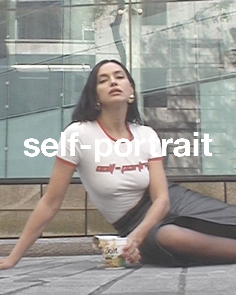 Portrait of a woman seated outdoors in a casual outfit, holding a cup, with a modern building visible in the background.