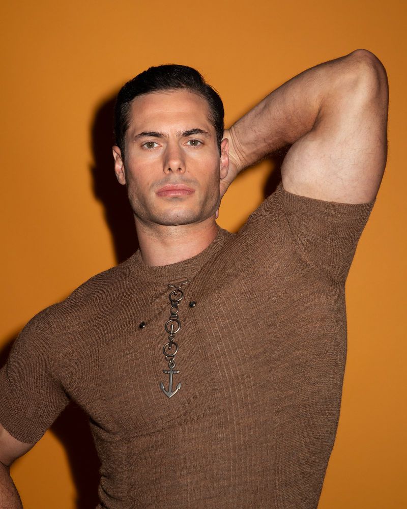 Attractive adult man with dark hair poses with arm up on an orange background wearing a brown knit shirt with an anchor pendant.