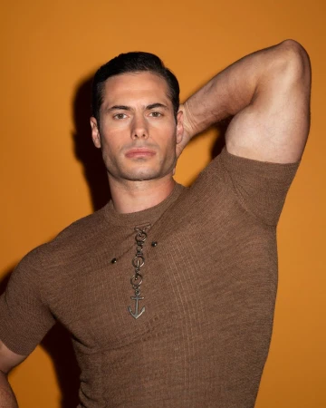 Attractive adult man with dark hair poses with arm up on an orange background wearing a brown knit shirt with an anchor pendant.