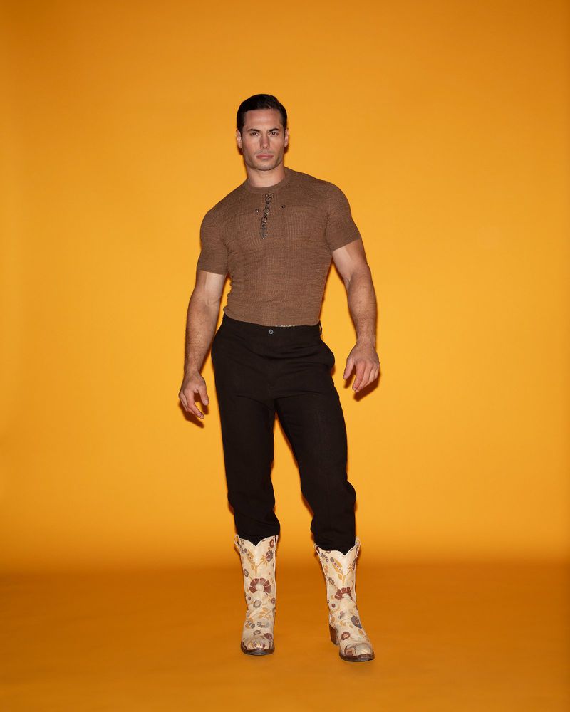 Masculine male model in brown short-sleeve shirt and black pants poses in decorated cowboy boots against an orange backdrop.