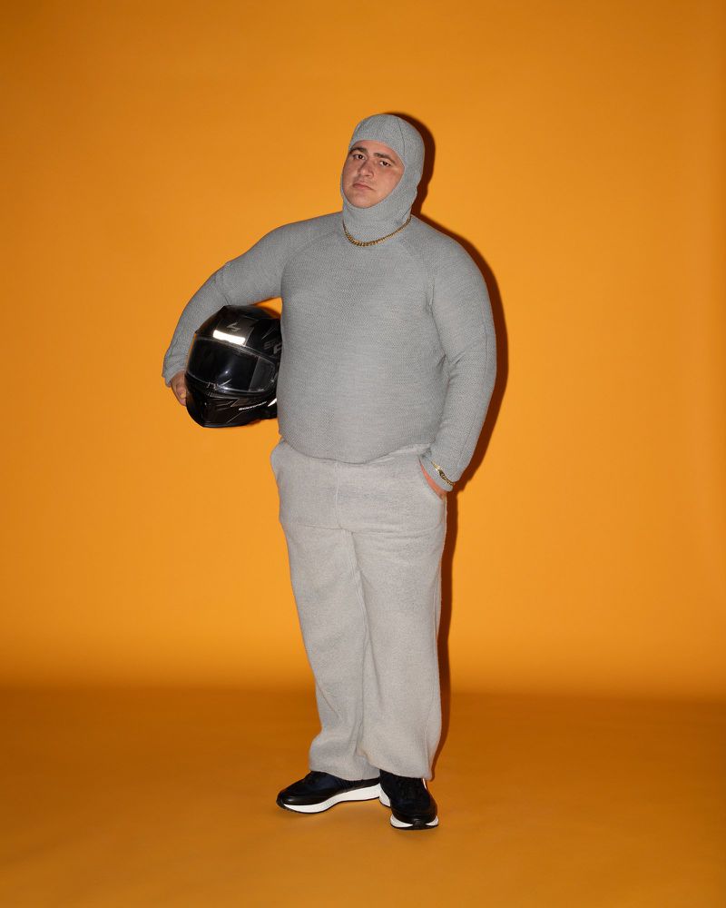 Adult man in a grey balaclava and sweatshirt holding a black motorcycle helmet against a bright orange background in a studio shot.