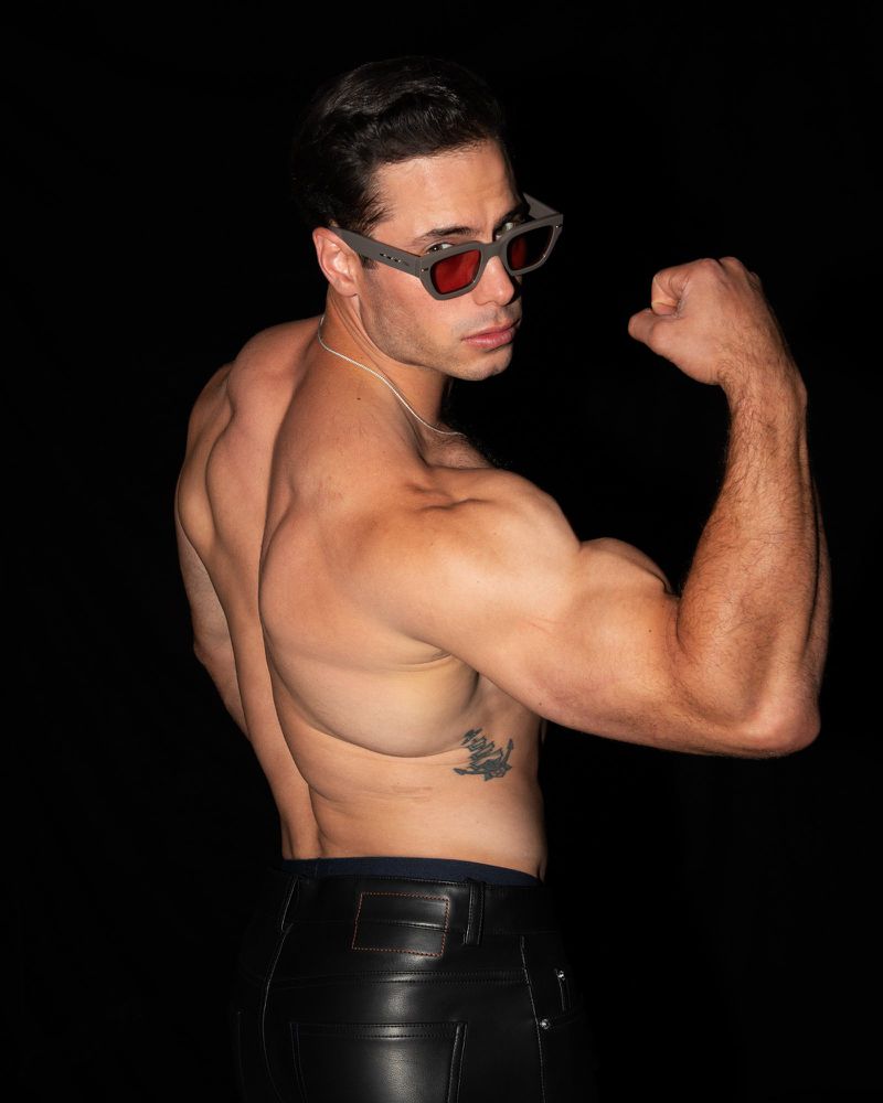 Portrait of shirtless muscular man flexing his biceps, wearing sunglasses and black leather pants against a dark background.