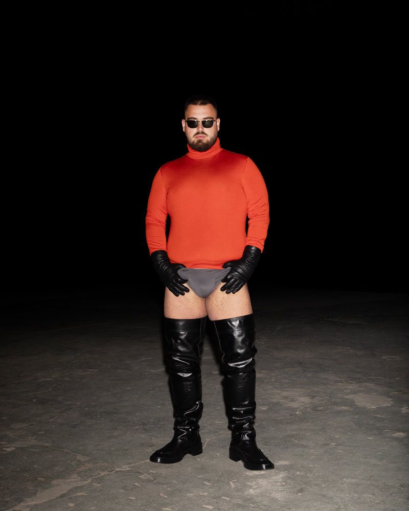 An adult male stands in a studio wearing a red turtleneck, black leather boots, black leather gloves, and minimal grey underwear.