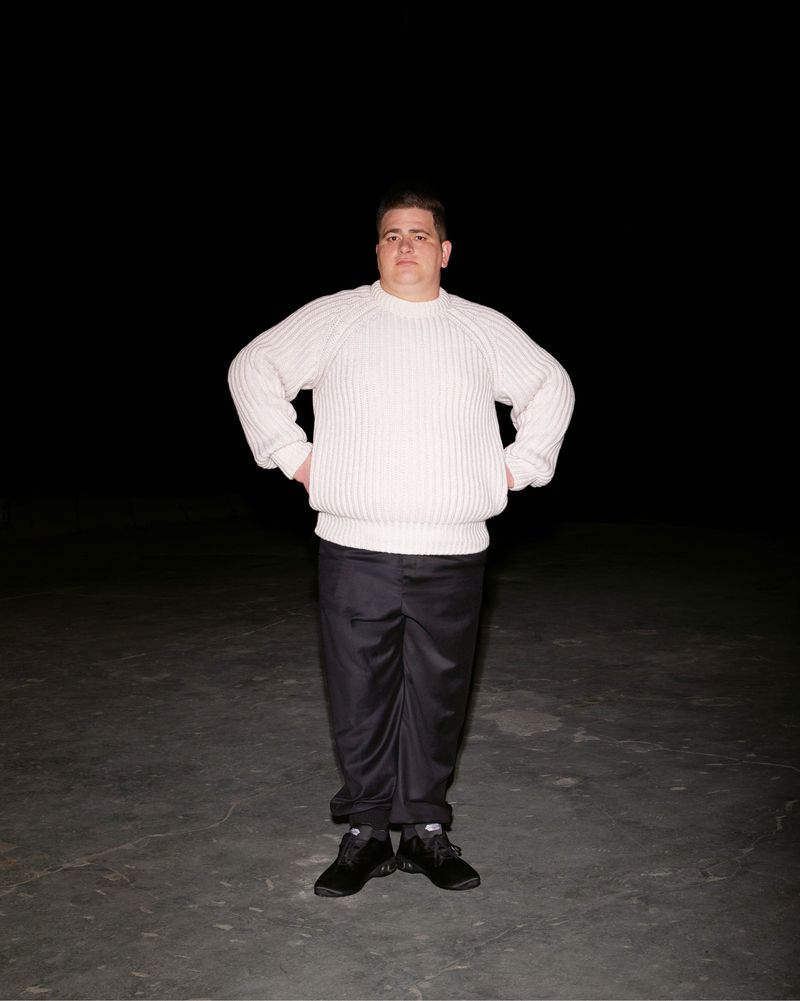 Full-length shot of an adult man in a cream sweater and black pants standing indoors against a dark background with his hands on his hips.