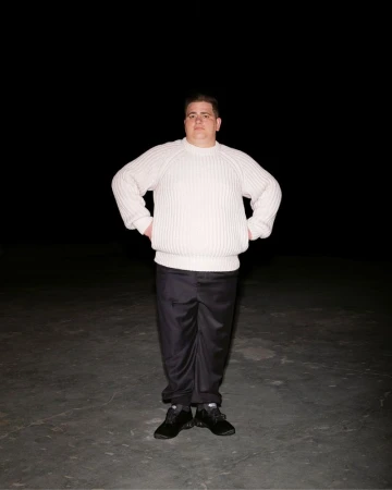 Full-length shot of an adult man in a cream sweater and black pants standing indoors against a dark background with his hands on his hips.