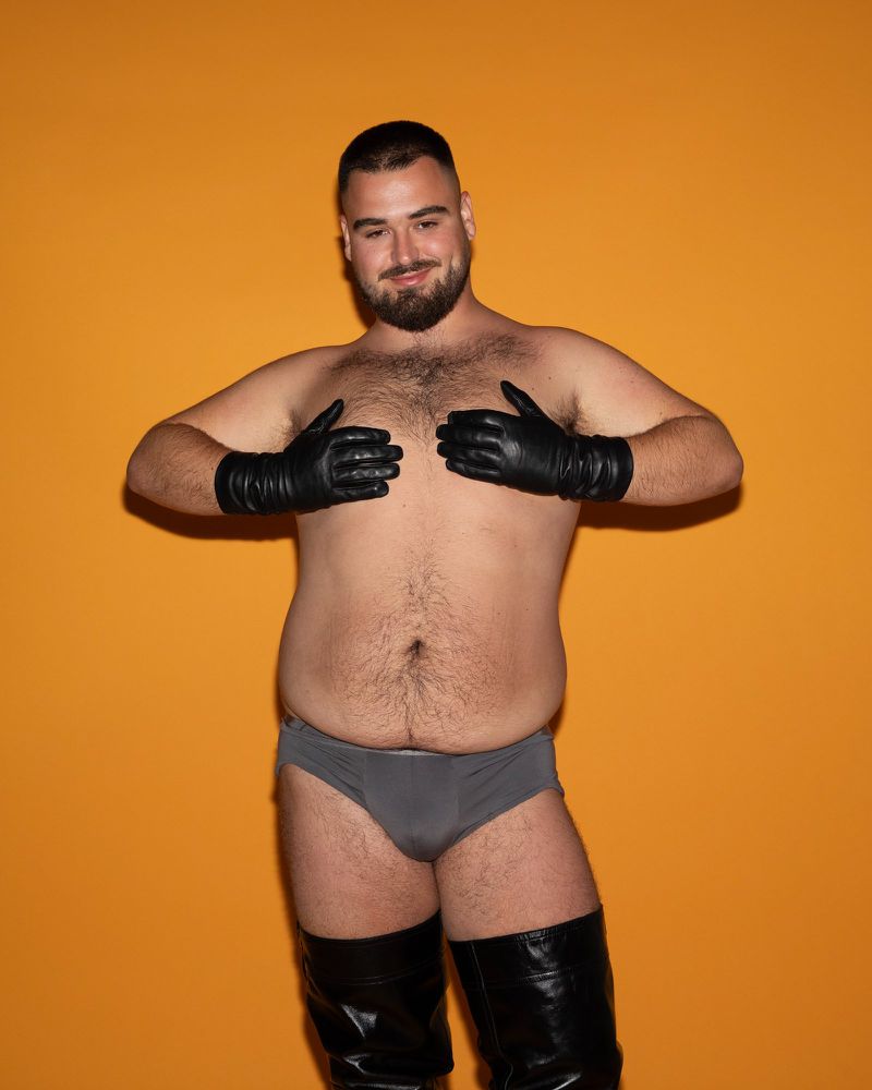 Adult male posing with gloves and boots against an orange backdrop, showcasing a fashion-forward and confident aesthetic.