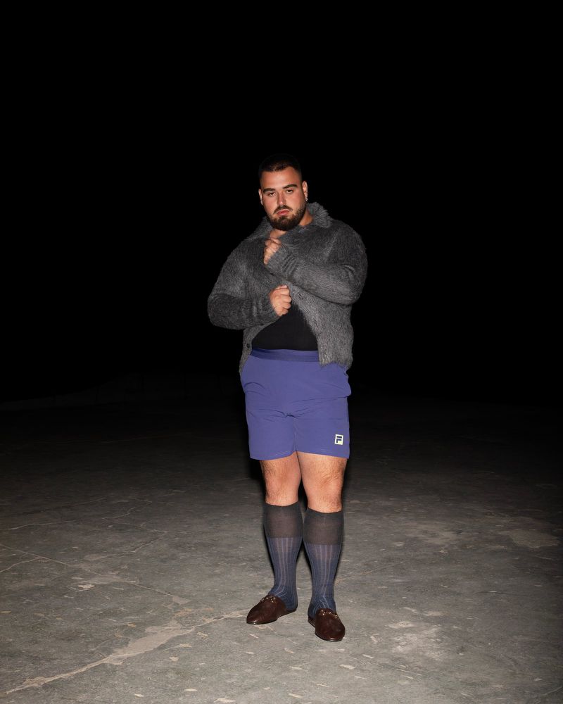 Adult male with beard and mustache stands in gray sweater, blue shorts, and dark socks and loafers on a concrete surface at night.
