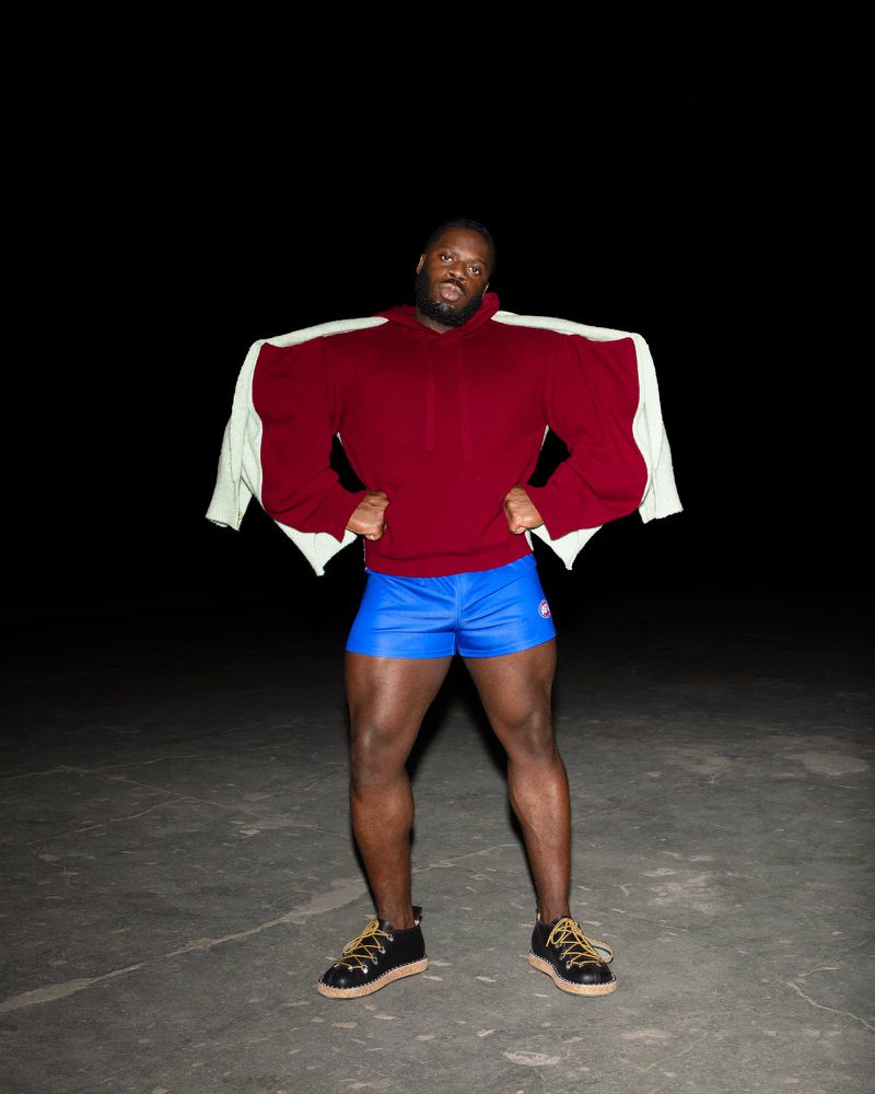 Man poses in a maroon hoodie, blue shorts and black shoes. There is a white jacket over his shoulders, set against a dark background.