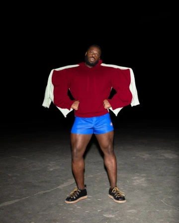 Man poses in a maroon hoodie, blue shorts and black shoes. There is a white jacket over his shoulders, set against a dark background.