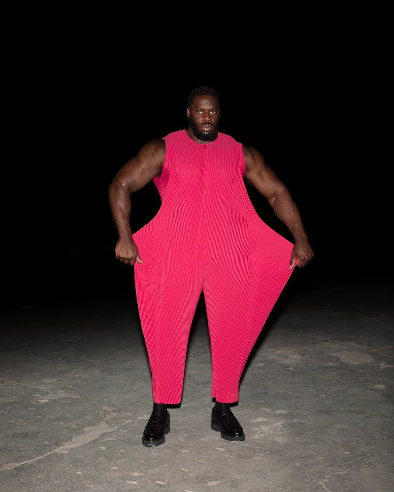 A muscular adult male in a red jumpsuit stands in a dark, outdoor setting. The clothing is oversized, and the lighting is studio-like.