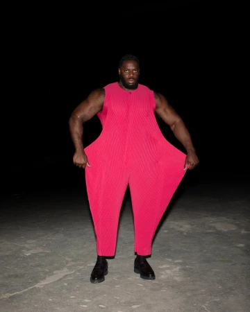 A muscular adult male in a red jumpsuit stands in a dark, outdoor setting. The clothing is oversized, and the lighting is studio-like.